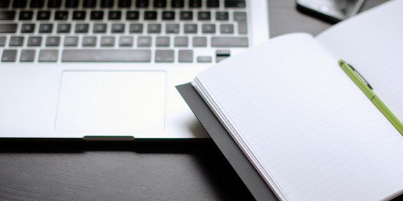 Laptop und Notizbuch mit Stift auf einem Tisch liegend.