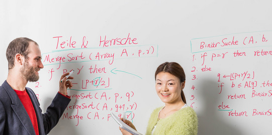 Lehrsituation-Whiteboard Zwei Personen stehen vor einem Whiteboard mit Formeln und Beschriftung. Eine Person hat einen Stift in der Hand, die andere einen Block.